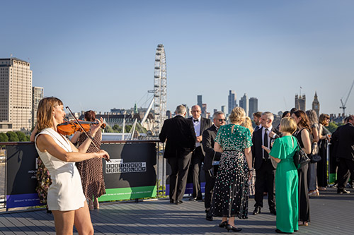 People starting to gather on the balcony of the Institute of Engineering and Technology for the National Innovation Challenge 2025 Awards evening. They are all dressed in formal wear and there is a violinist playing in the foreground with the London skyline behind them.