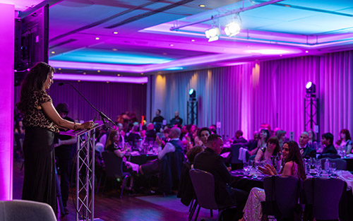 Konnie Huq is on stage, in front of an audience, speaking at the 2025 National Innovation Challenge Awards night. She is standing behind a lectern and the audience are seated around tables.
