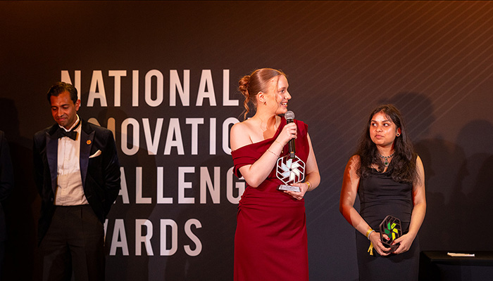 Two 2026 National Innovation Challenge female finalists on stage at the awards ceremony. Dressed in formal wear, holding their trophies. One of them is wearing a red dress and holding a microphone and there is a judge to their left dressed in a tuxedo. The words: National Innovation Challenge Awards are behind them.