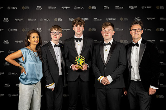Keith Smith, CEO of HRUC and BBC presenter and STEM ambassador Konnie Huq posing with the schools category winners (from St Bede’s Catholic High School) of the National Innovation Challenge 2026. All wearing formal attire standing in front of a wall of logos.