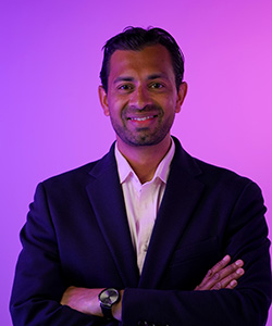 Sarwar Khan standing with his arms folded and smiling. He is wearing a dark navy jacket and white shirt and standing in front of a pink background.
