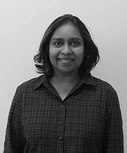 Thiloshini Ellary-Bachu is wearing a checked shirt and smiling. She is standing in front of a plain white background.   