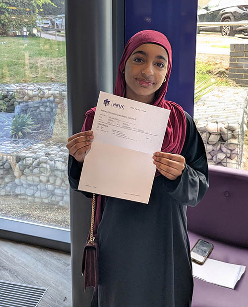 Female Harrow College student holding up her GCSE results letter in front of a purple pillar with windows behind her.