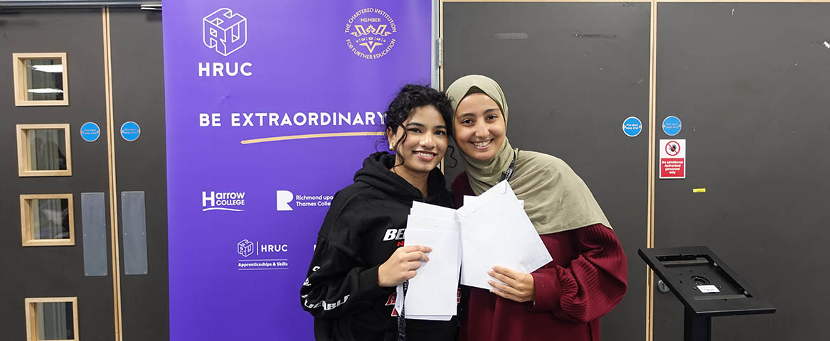 Two female Uxbridge students holding their GCSE results envelopes. They are smiling at the camera and standing in front of a Be Extraordinary purple banner with HRUC logo.   