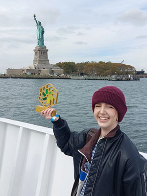 Emily Cheesman - one of the National Innovation Challenge winners holding her trophy up, with the Statue of Liberty behind her. 