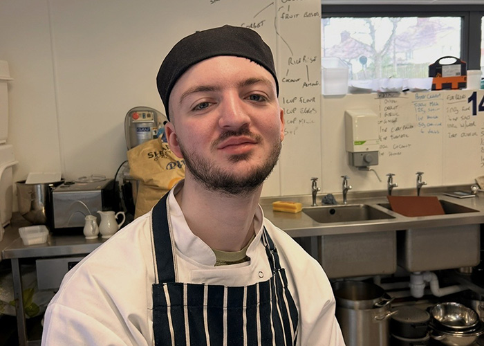 Alfie Green - alumni student from Richmond upon Thames College (RuTC) wearing his chef uniform, stripe apron and hat, sitting in a commercial kitchen setting. There are sinks behind him. 