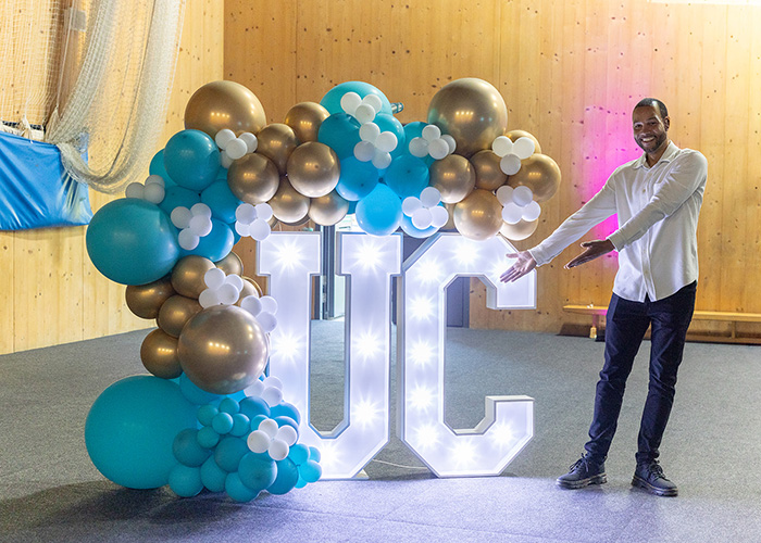 Anthony Bennett is standing next to a lit up UC sign with blue, white and gold balloons on top. He is gesturing towards the sign.  