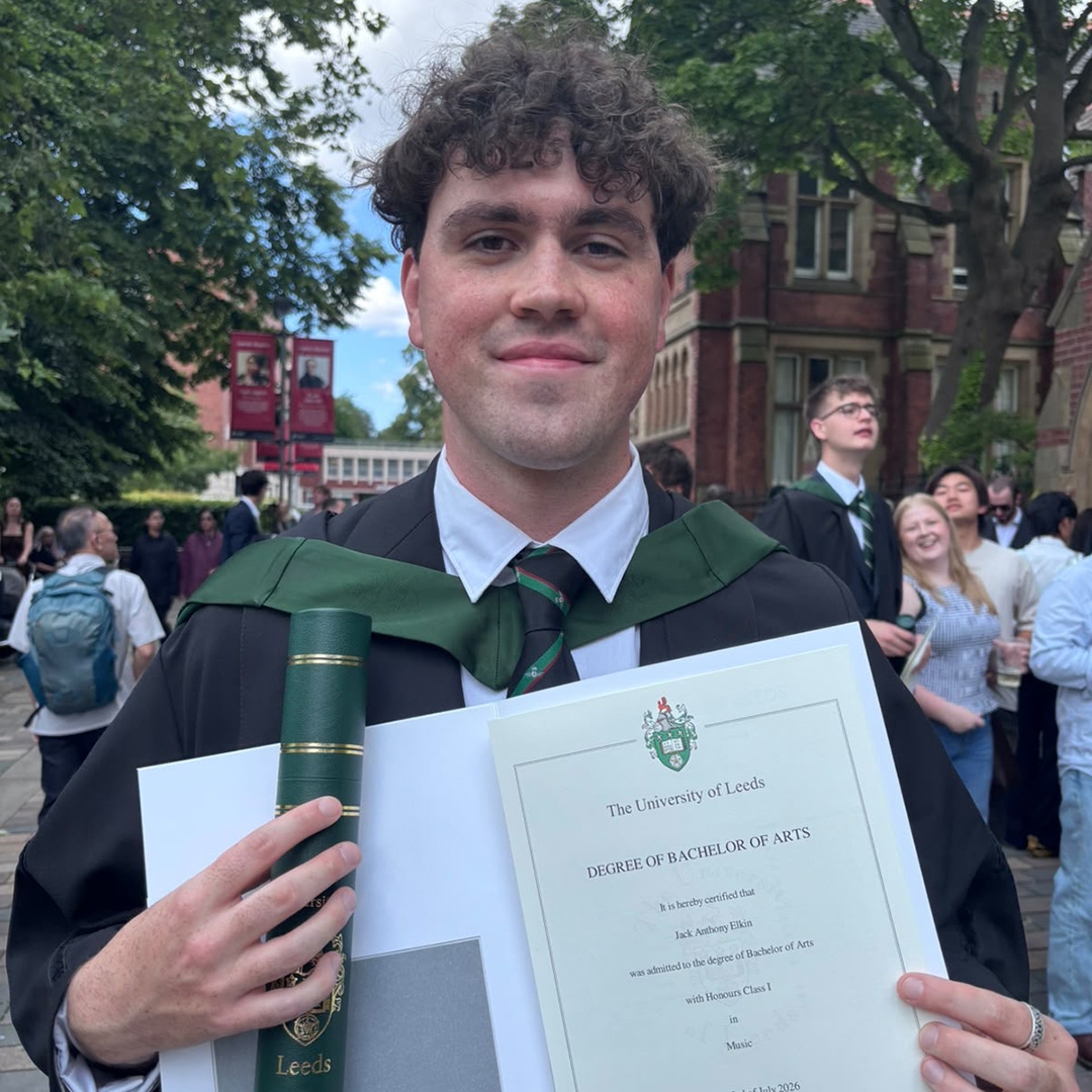 Jack Elkin - alumni - graduate of the Music Production and Performance UAL Level 3 Extended Diploma at RuTC from 2020 to 2022. He is wearing his graduation robes with a green sash and is holding his degree proudly. There are other students in the background with trees around them.