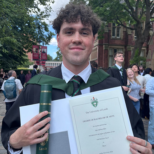 Jack Elkin - alumni - graduate of the Music Production and Performance UAL Level 3 Extended Diploma at RuTC from 2020 to 2022. He is wearing his graduation robes with a green sash and is holding his degree proudly. There are other students in the background with trees around them.