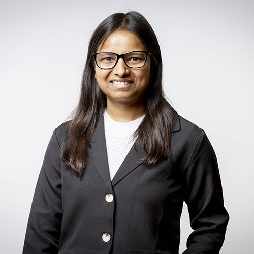 Pragna Navinkumar - former Harrow College student. She is wearing a black jacket and white top and has glasses with long brown hair.