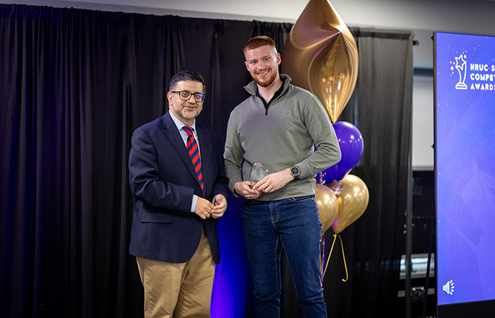Samuel Stevely receiving his Skills Award and standing with Asif Khan - Assistant Principal at Uxbridge College on stage. There is a black curtian with purple and gold balloons behind them. 