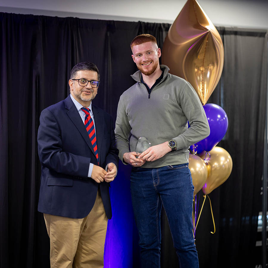 Samuel Stevely receiving his Skills Award and standing with Asif Khan - Assistant Principal at Uxbridge College on stage. There is a black curtian with purple and gold balloons behind them. 