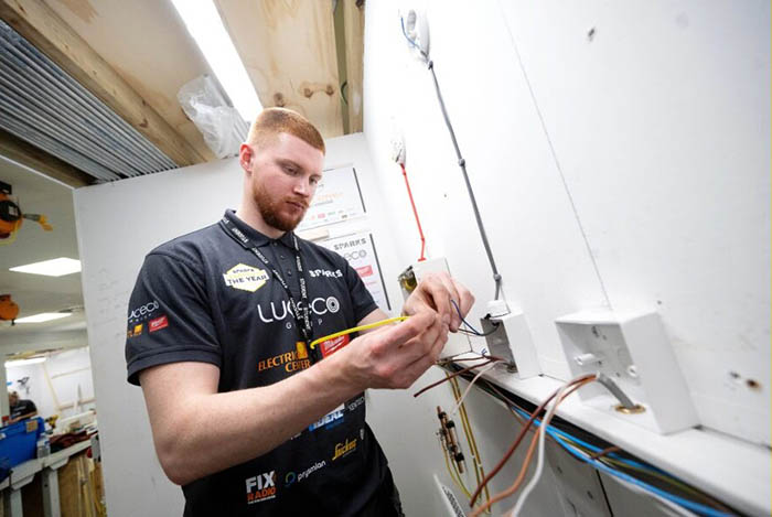 Samuel Stevely practising electrical installation in a workshop. He is wearing a black branded t-shirt with numerous logos.