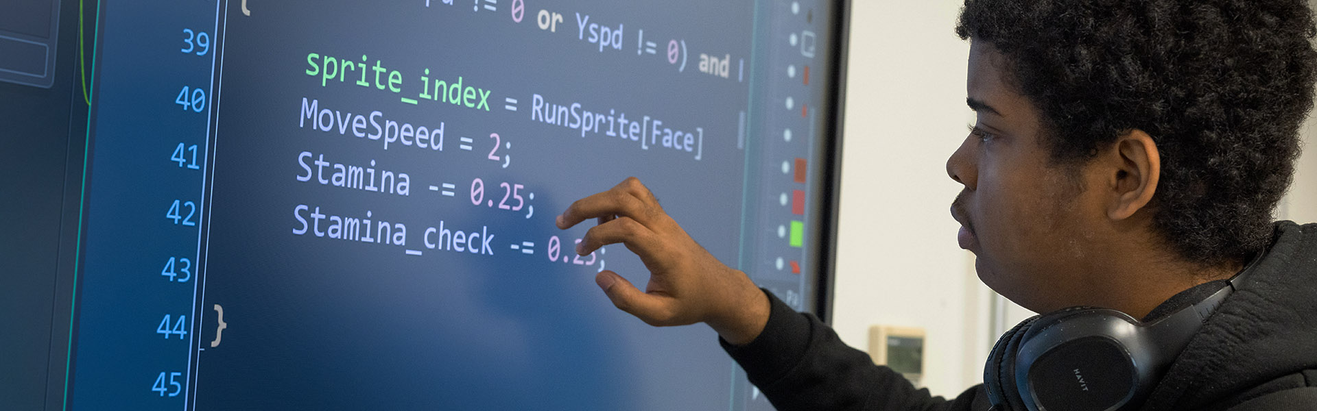 A male Uxbridge student working on an IT problem on an interactive screen. He is wearing black headphones around is neck, a black hoodie and touching the blue screen.  