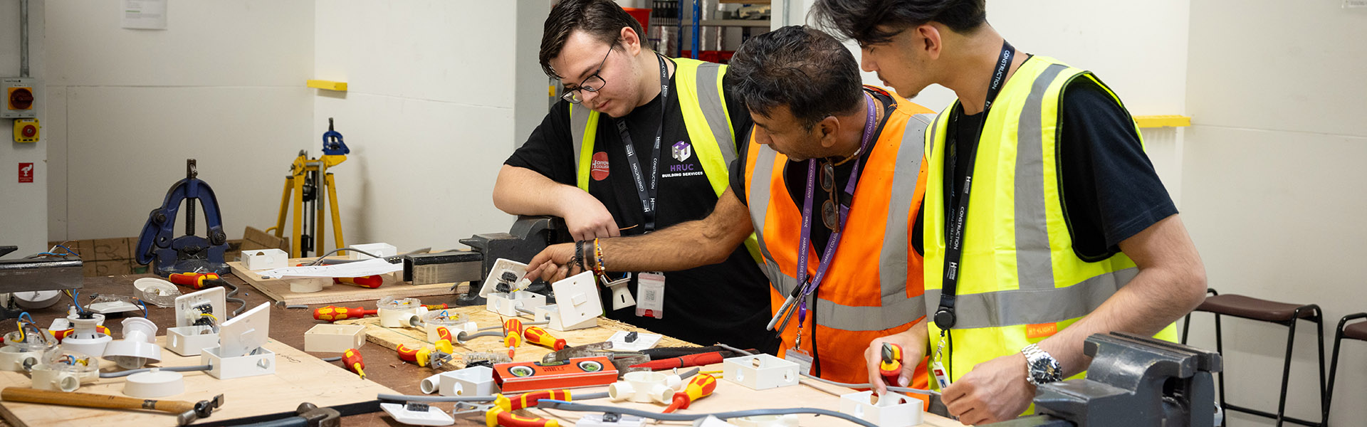 Harrow College teacher showing Construction students how to do electrical installation techniques on electrical switches. They have tools and switches across a workbench.