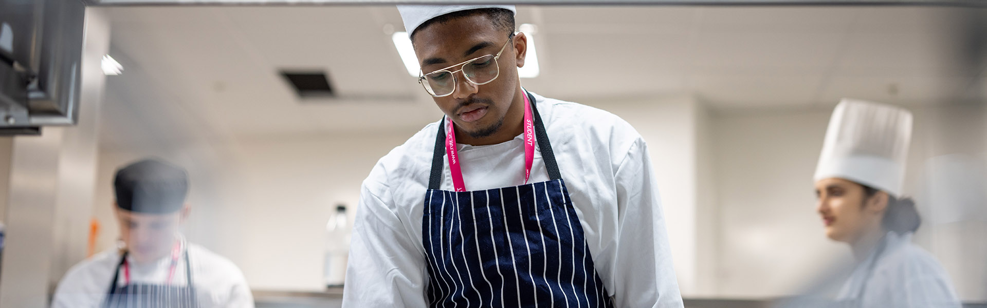 Richmond upon Thames College students cooking in The View Restaurant kitchen - they are wearing white chef hats and blue and white striped aprons. 