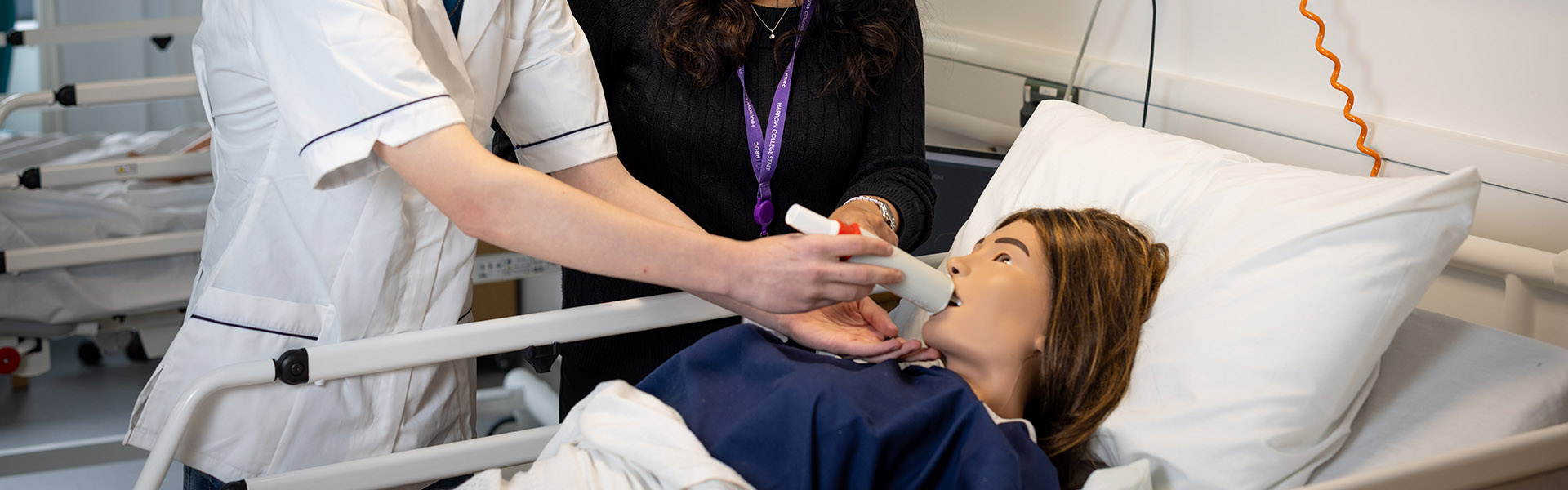 Harrow College Health and Social Care student practising taking care of a patient using a manikin which is lying in a hospital bed. A tutor is watching his technique of how he is administering something by mouth.