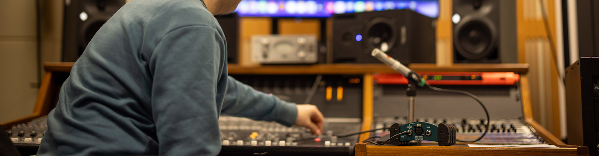 Male Richmond upon Thames music student in the studio at the sound desk, adjusting one of the inputs. There is a microphone next to him.   