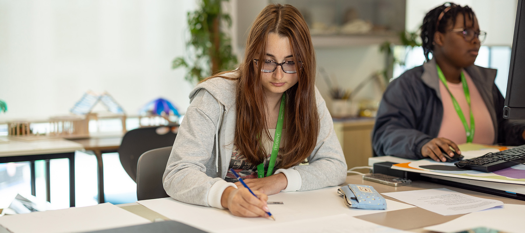 Female Richmond upon Thames College student in class and and writing on paper. She is concentrating on what she is doing. There is another female student next to her on a computer. 