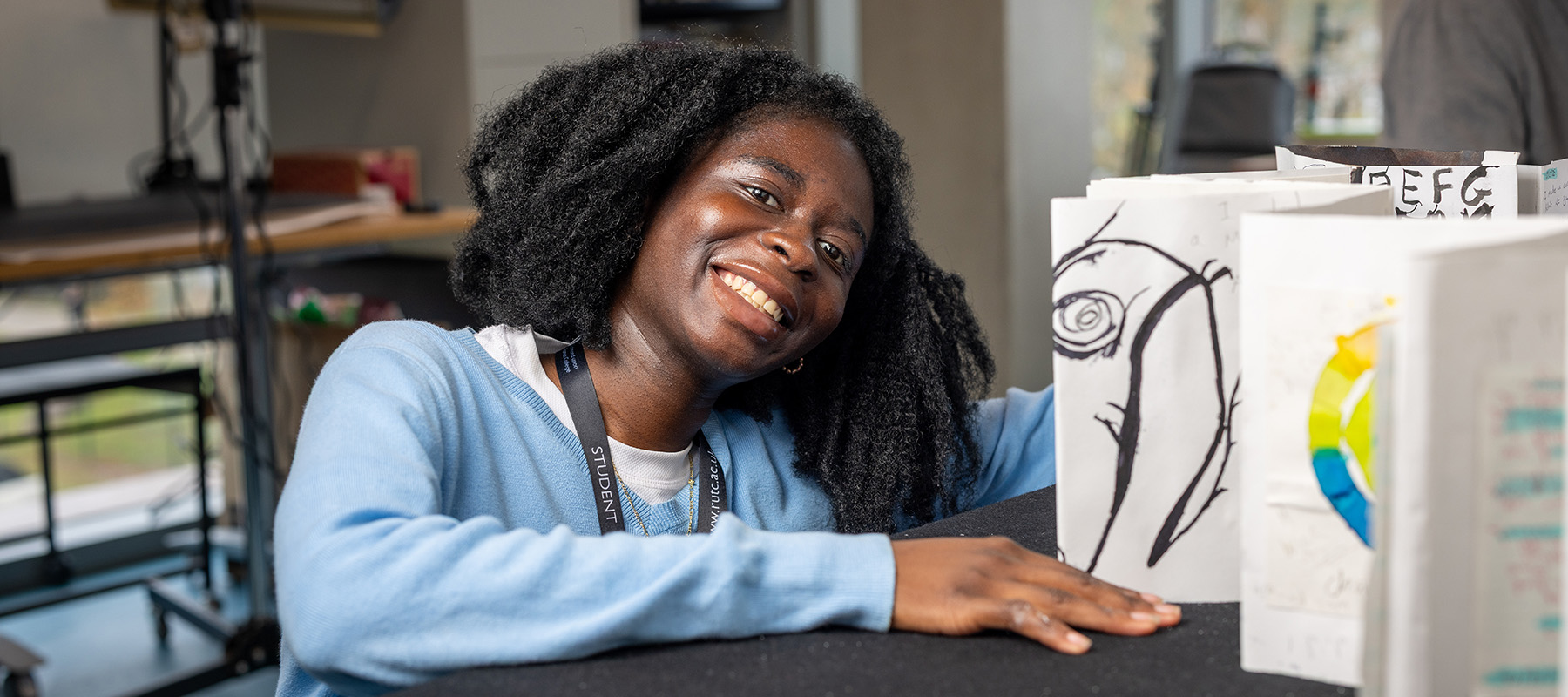 Richmond upon Thames College art student sitting in front of her work on the table. She is in the workshop and is wearing a blue jumper and has dark neck length hair.