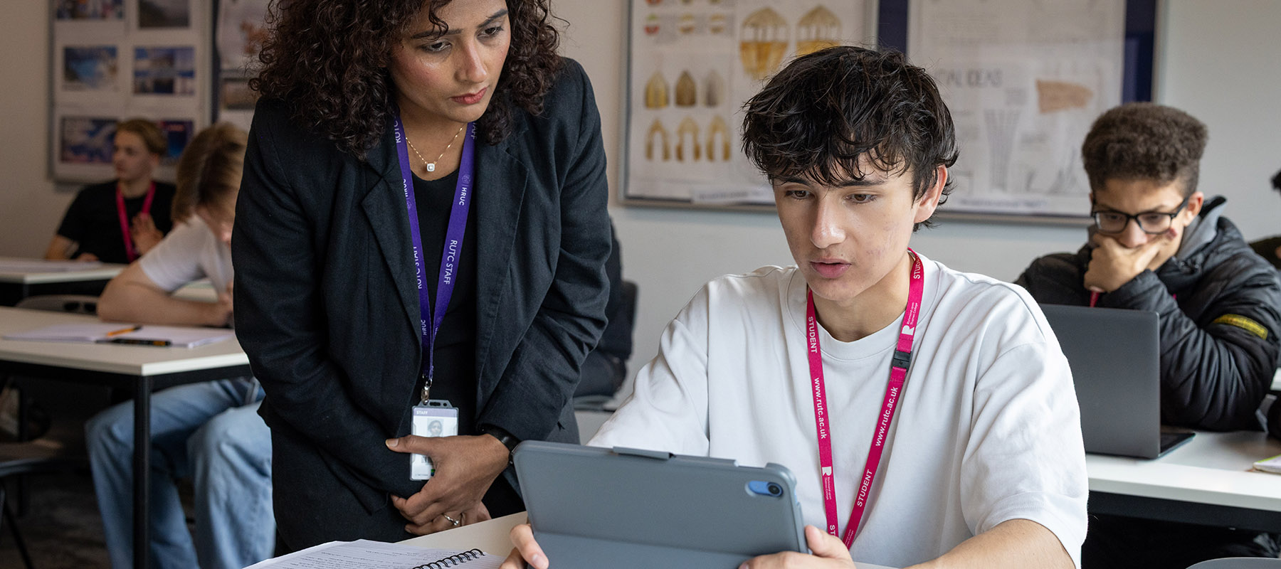 A female Richmond upon Thames teacher helping a male student in class. They are looking at his tablet, both are wearing lanyards.