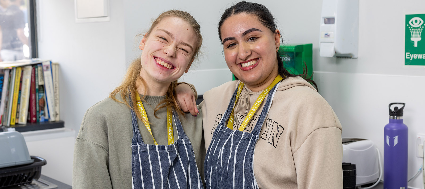 Two female Uxbridge College Inclusive Learning students standing in the College kitchen facilities, wearing blue and white stripe aprons and smiling.