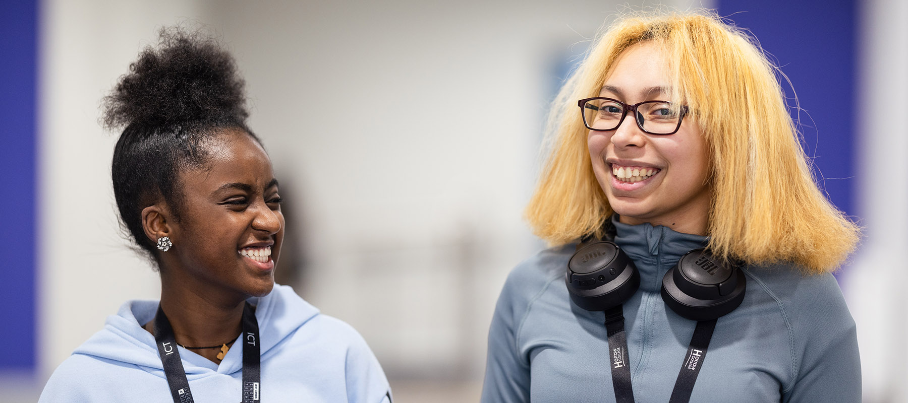 Two female ICT Harrow College students walking down the hall, smiling and laughing together. The girl on the left has her hair in a top knot and the girl on the right has bleached blond hair and wears glasses. 