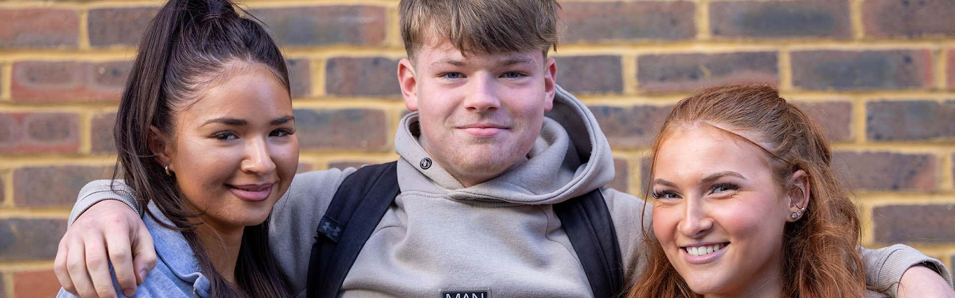 Three Uxbridge College students smiling at the camera. Two female students standing either side of one male student with his arms around their shoulders, in front of a brick wall. 