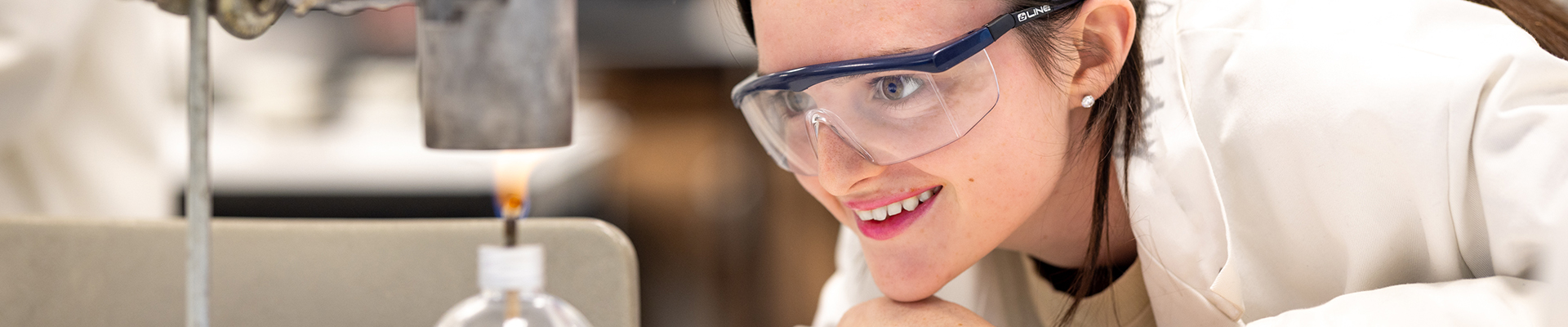 A Richmond upon Thames student in biomedical science conducting an experiment in a lab. She is heating a beaker and watching the results, wearing safety glasses and a white lab coat.  