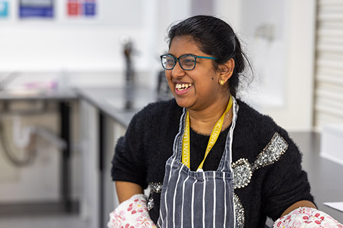 Female Uxbridge College Foundation Learning student cooking in the kitchen facilities. She is smiling, wearing a blue and white striped apron and a flowery oven mit.