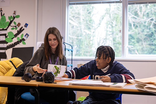Harrow Weald Foudation Learning Student working with a female staff member. They are holding a pen over a work book and looking at another book with drawings. Both are sitting at a desk.