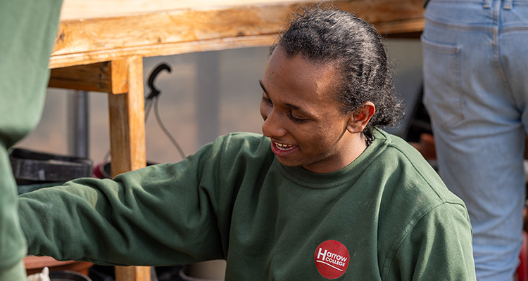 Harrow Weald Campus Foundation Learning student working in the dedicated outdoor area as part of the Spring House centre. He and other students are potting soil into multi-plant pot trays. He is wearing a green jumper with the Harrow College red logo.