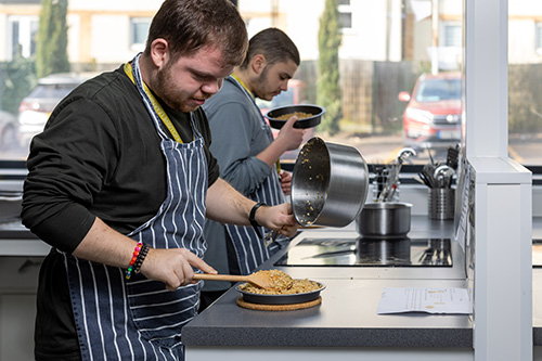 2 x male Uxbridge College Foundation Learning students baking in the kitchen facilities. They are both wearing striped aprons. The student closest is spooning a mixture into a round baking tray.