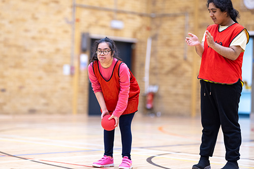 2 x Richmond upon Thames Foundation Learning students playing with an orange ball in a sports hall. They are wearing orange vests.