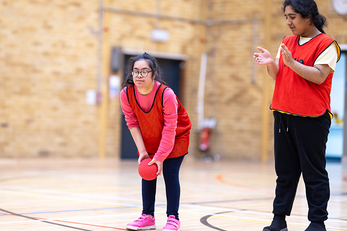 2 x Richmond upon Thames Foundation Learning students playing with an orange ball in a sports hall. They are wearing orange vests. 