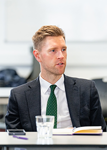 John Cope the new Chair of HRUC's Board of Governors in a grey suit and green with white dots tie. He is sitting at a table in mid-conversation. There is a glass, pen, notebook and mobile on the table in front of him.