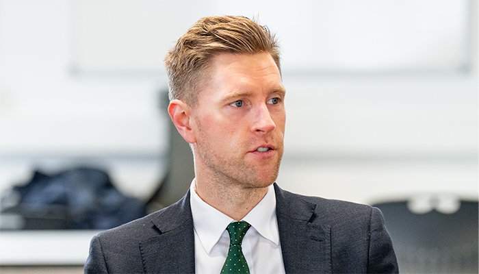 John Cope the new Chair of HRUC's Board of Governors in a grey suit and green with white dots tie. He is sitting at a table in mid-conversation. There is a glass, pen, notebook and mobile on the table in front of him.  
