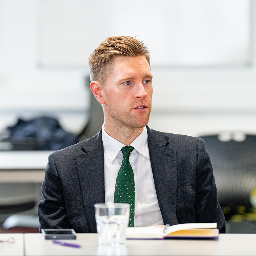 John Cope the new Chair of HRUC's Board of Governors in a grey suit and green with white dots tie. He is sitting at a table in mid-conversation. There is a glass, pen, notebook and mobile on the table in front of him.  