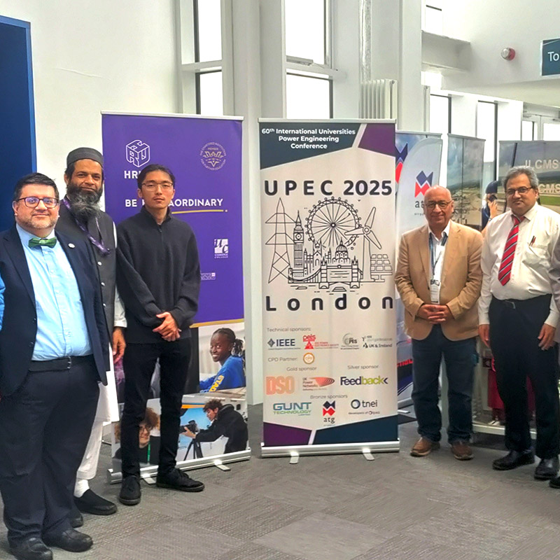 Bektur Ogobaev (wearing glasses and dressed in all black standing in front of the purple banner) posing for a group photo with other members of HRUC, Brunel University and delegates from the Universities Power Engineering Conference (UPEC 2025).