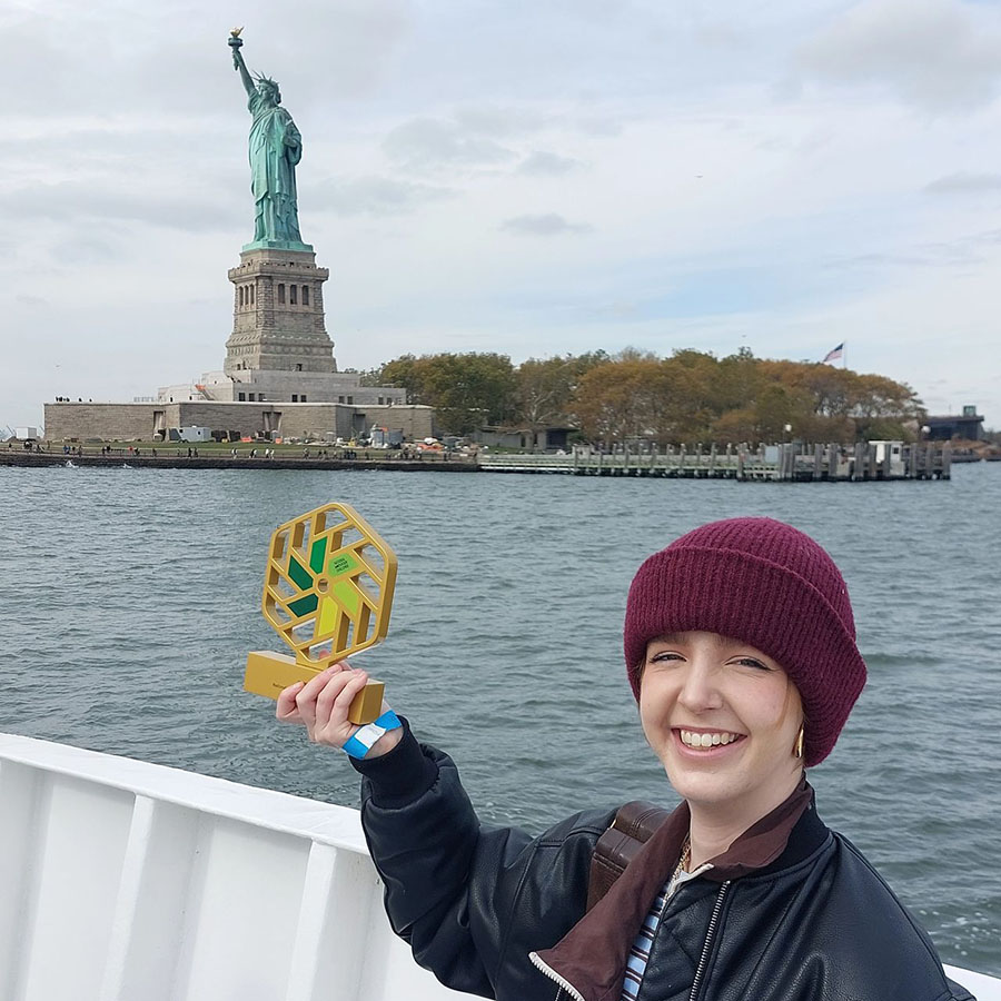 Emily Cheesman - one of the National Innovation Challenge winners holding her trophy up, with the Statue of Liberty behind her.