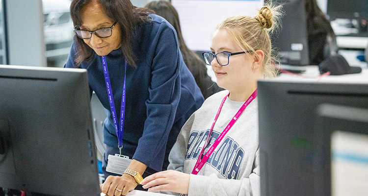 An HRUC student and staff member looking at a computer screen, both are wearing glasses. There are students working at computers in the background.