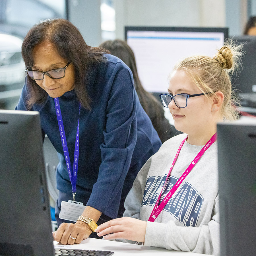 An HRUC student and staff member looking at a computer screen, both are wearing glasses. There are students working at computers in the background.