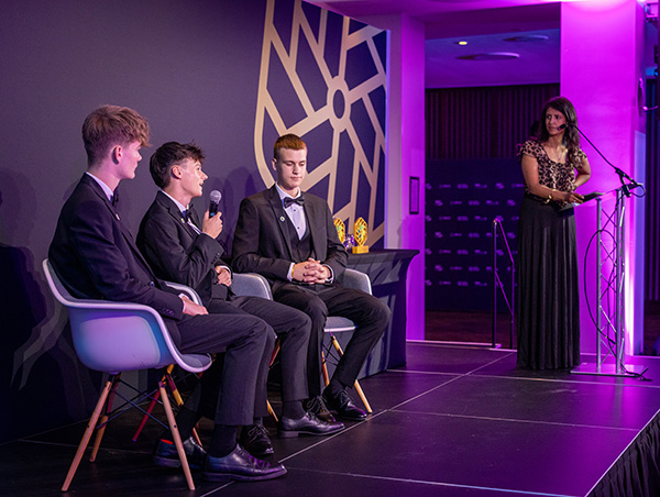 Students on stage with Konnie Huq at the 2025 National Innovation Challenge Awards night. The students are sitting and one is speaking into a microphone. They are all wearing tuxedos. Konnie is standing to the right at a lectern. The pinwheel branding of the NIC is on a backdrop behind them.