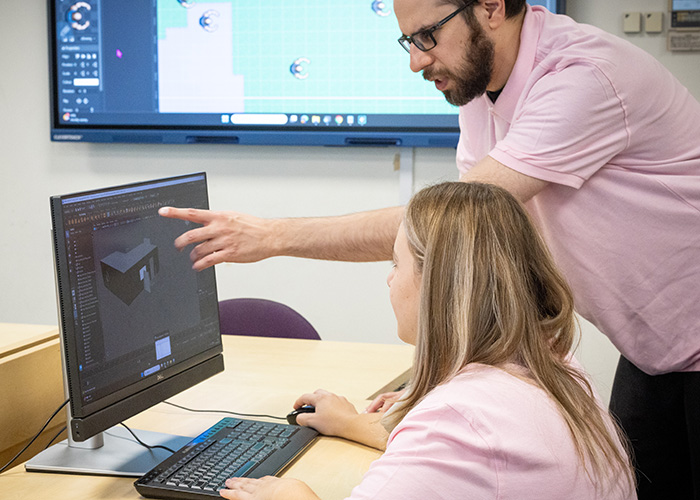 Male Richmond upon Thames College teacher showing a female student something on a computer screen. They are both wearing pink polo shirts and behnind them there is a large screen with a computer programme on it.