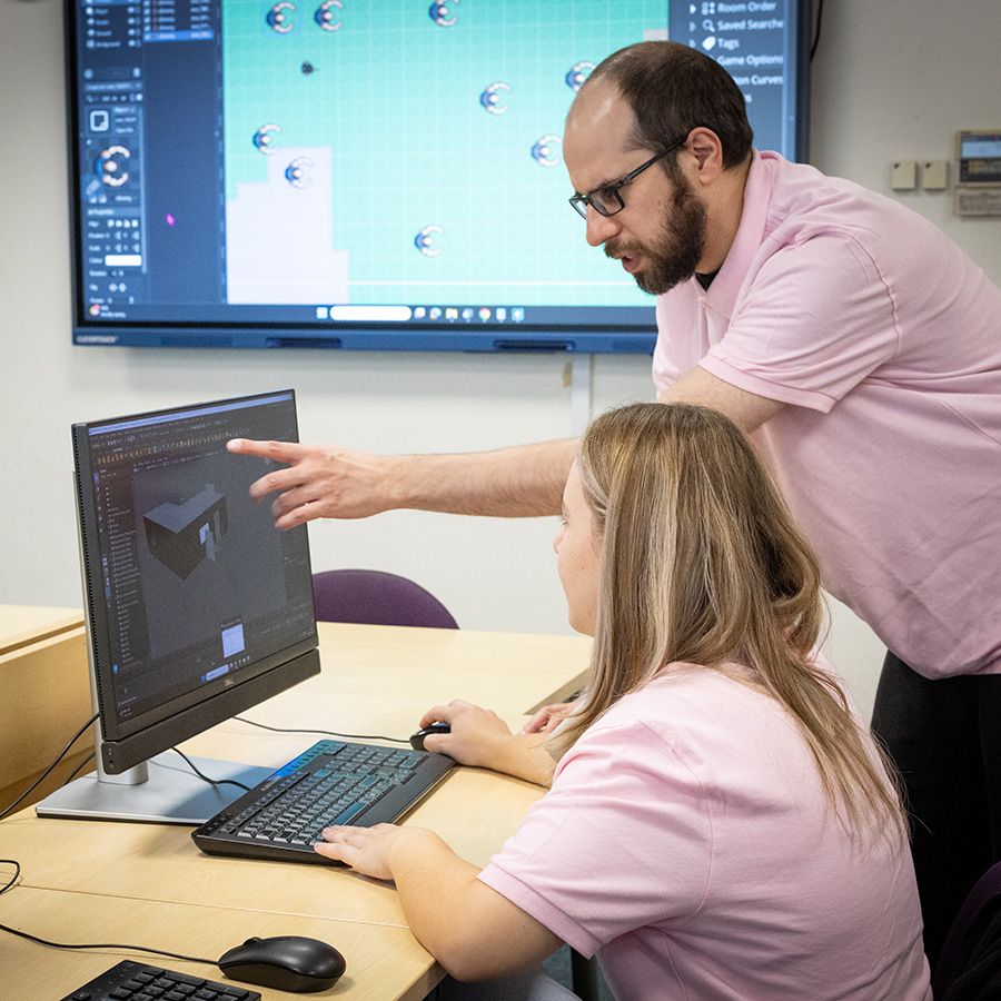 Male Richmond upon Thames College teacher showing a female student something on a computer screen. They are both wearing pink polo shirts and behnind them there is a large screen with a computer programme on it.