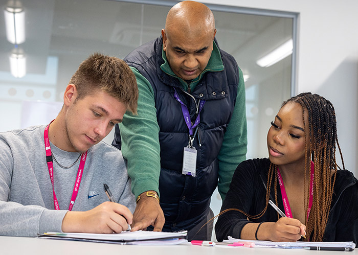 Richmond upon Thames College teacher helping two students. There is a male and female student sitting at a desk and looking at a notebook that the teacher is pointing to.