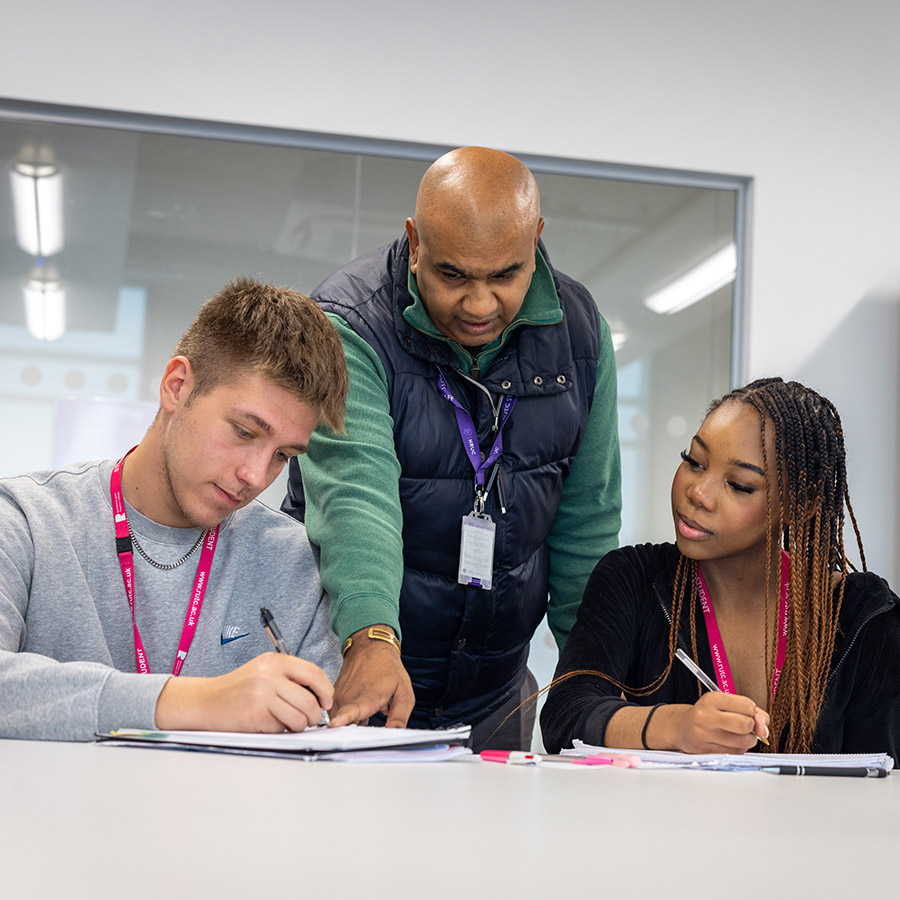 Richmond upon Thames College teacher helping two students. There is a male and female student sitting at a desk and looking at a notebook that the teacher is pointing to.