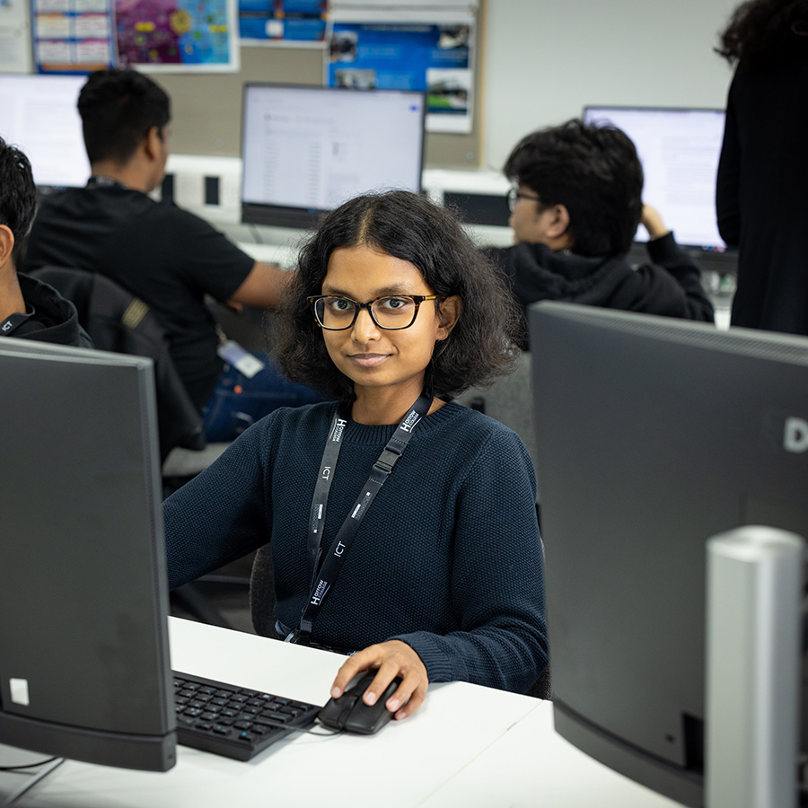 Female ICT Harrow College student sitting at a computer in a computer lab with other students around her working at their computers. There is a screen in the foreground and a keyboard and mouse in front of her. She is wearing glasses.