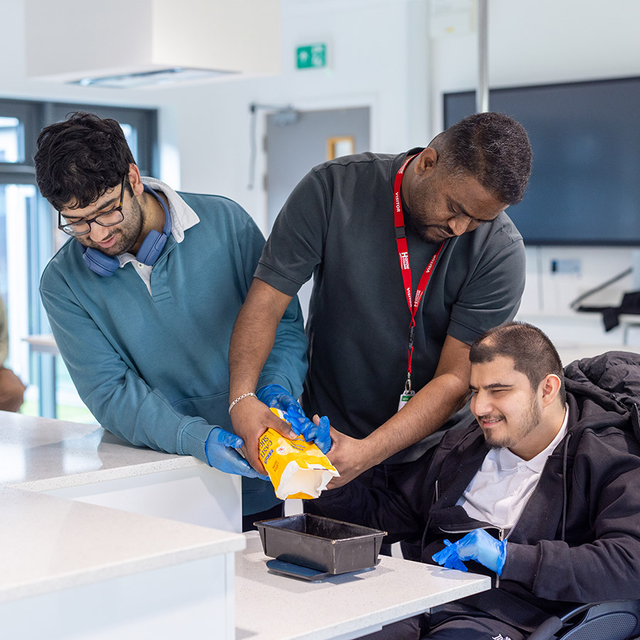 Foundation Learning students working in the Harrow Weald kitchen facilities, baking with a staff member. They are pouring caster sugar into a tin.