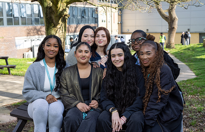 A group of female students sitting together in a group outside on a bench in the courtyard at Uxbridge College. They are smiling and there are trees and other students behind them. 